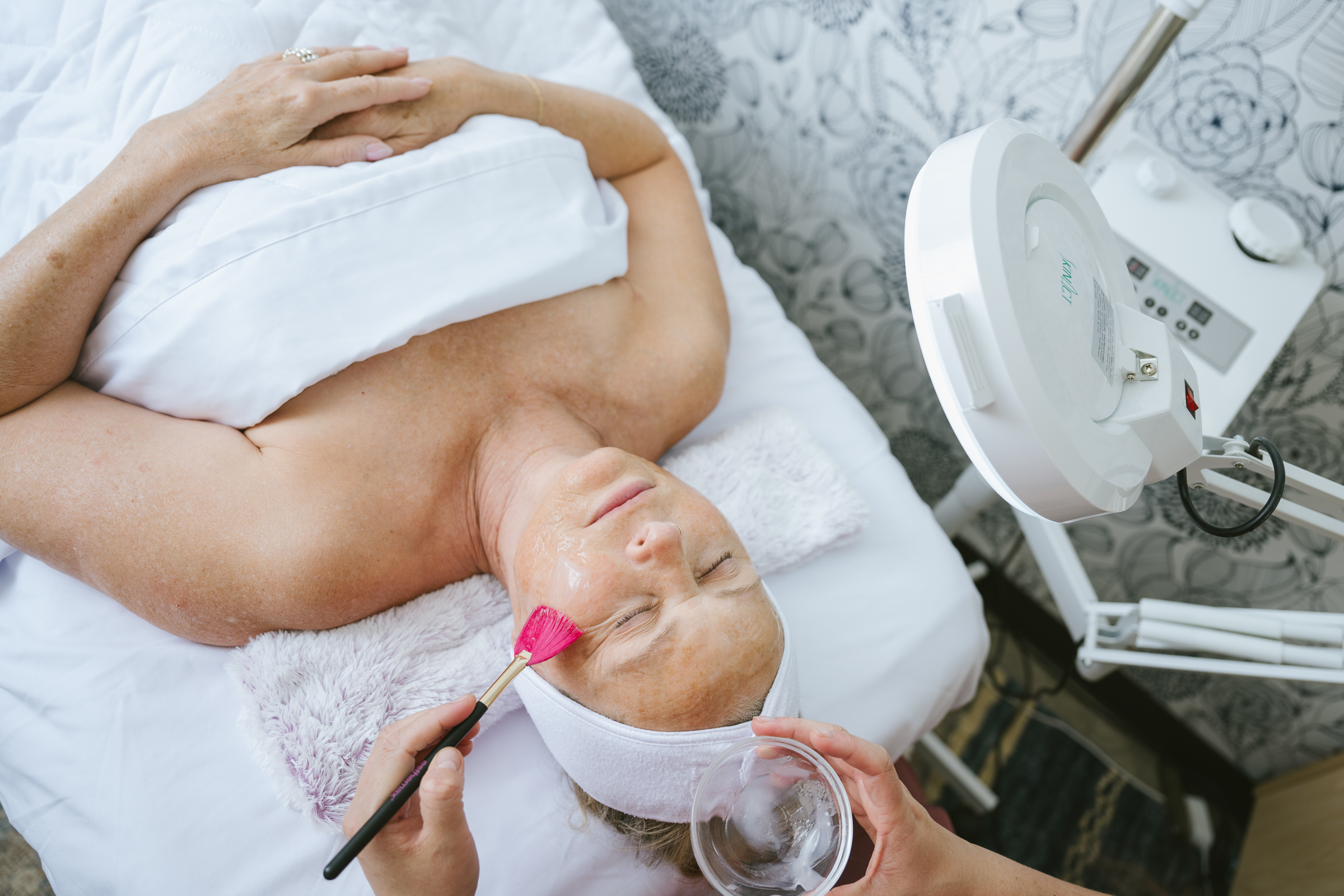 A woman receiving a chemical peel facial treatment at a spa, with an esthetician applying the solution using a brush.