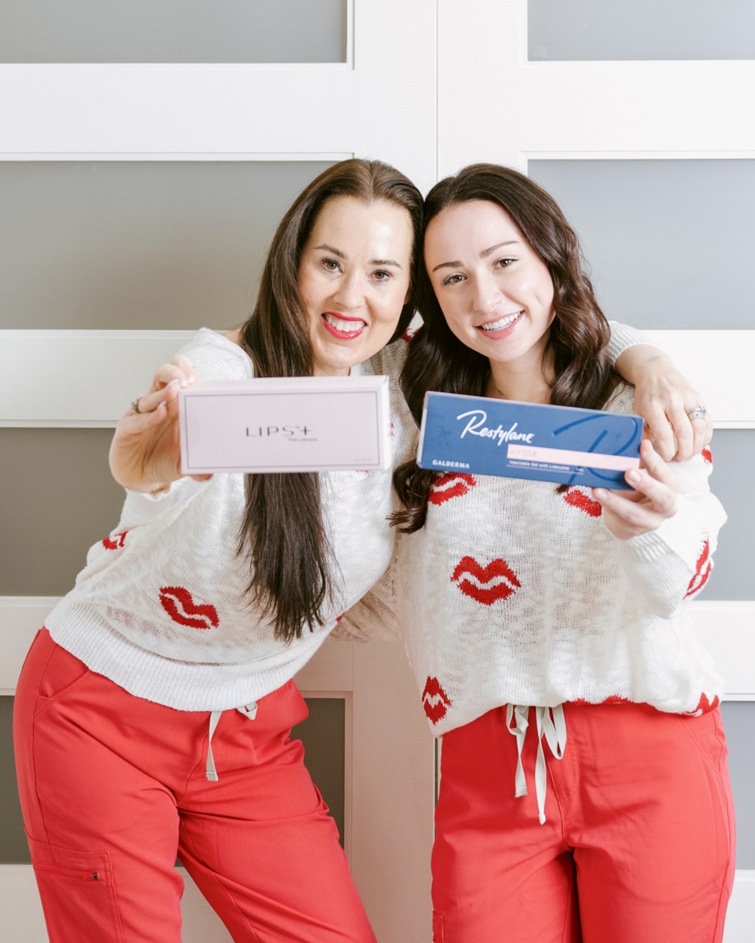 Two women standing side by side holding boxed skincare products while wearing matching outfits.