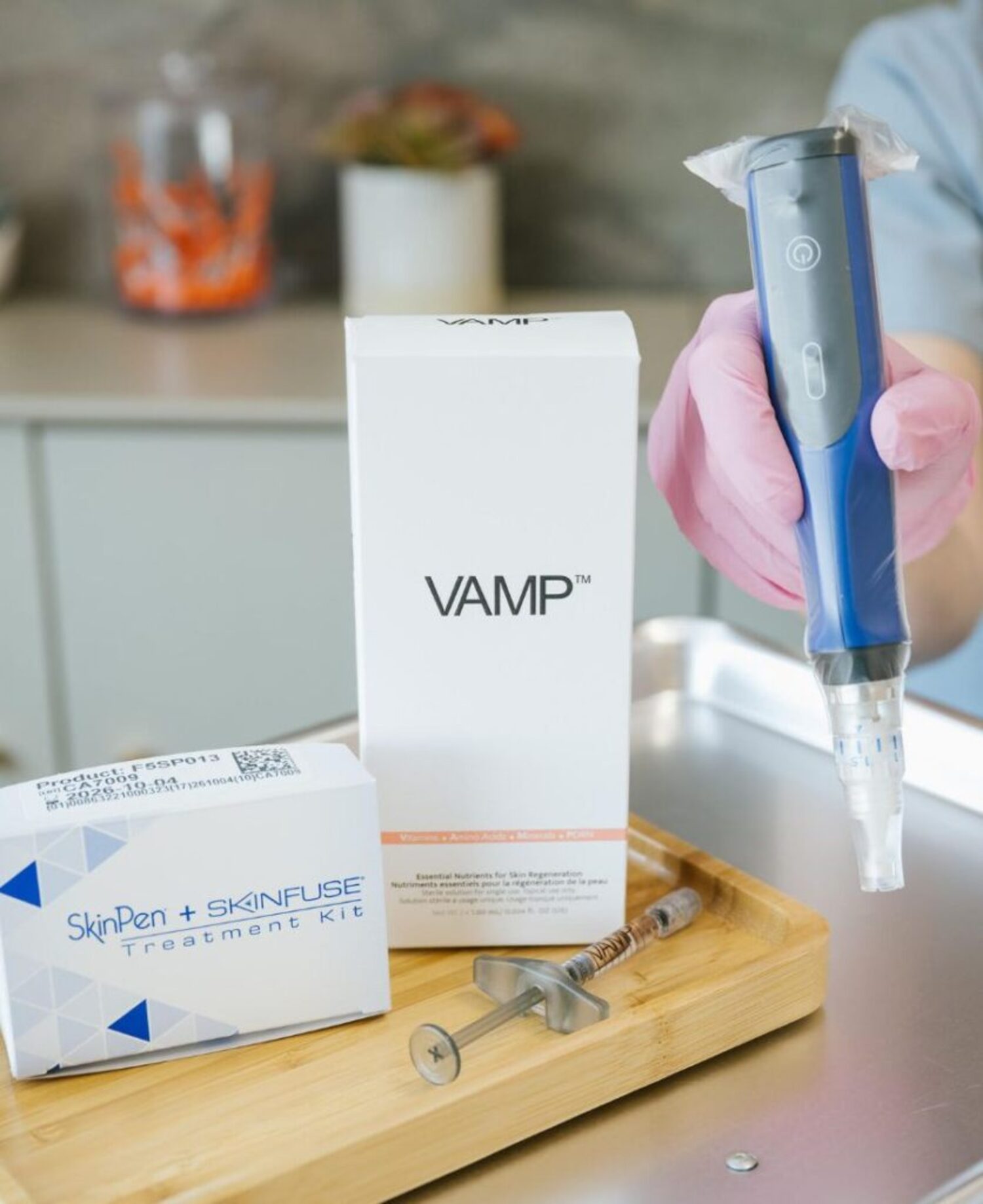 Gloved hand holding microneedling device next to boxed supplies, syringe, and tray on countertop.