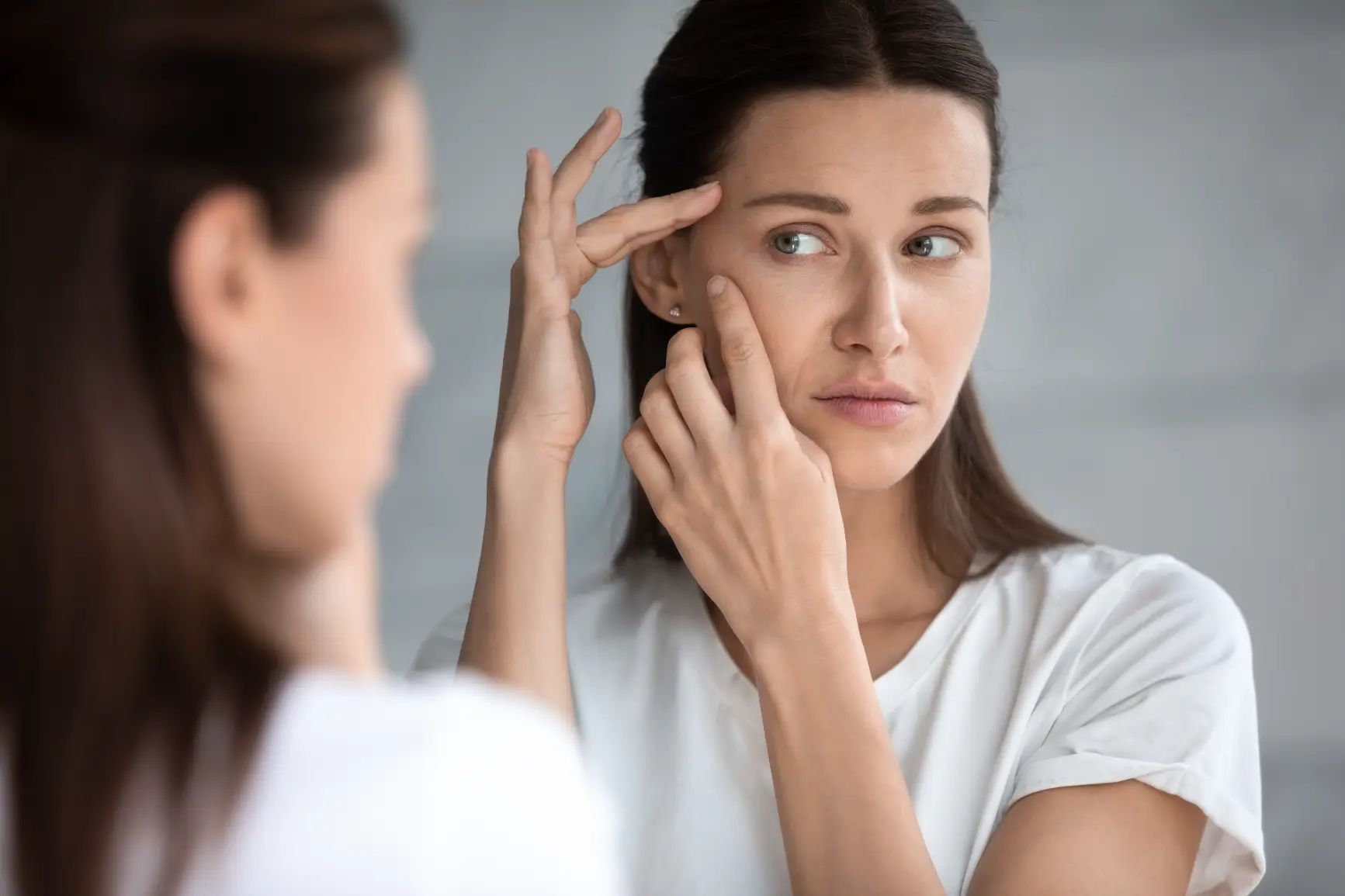 Woman examining facial lines near eye while looking in mirror