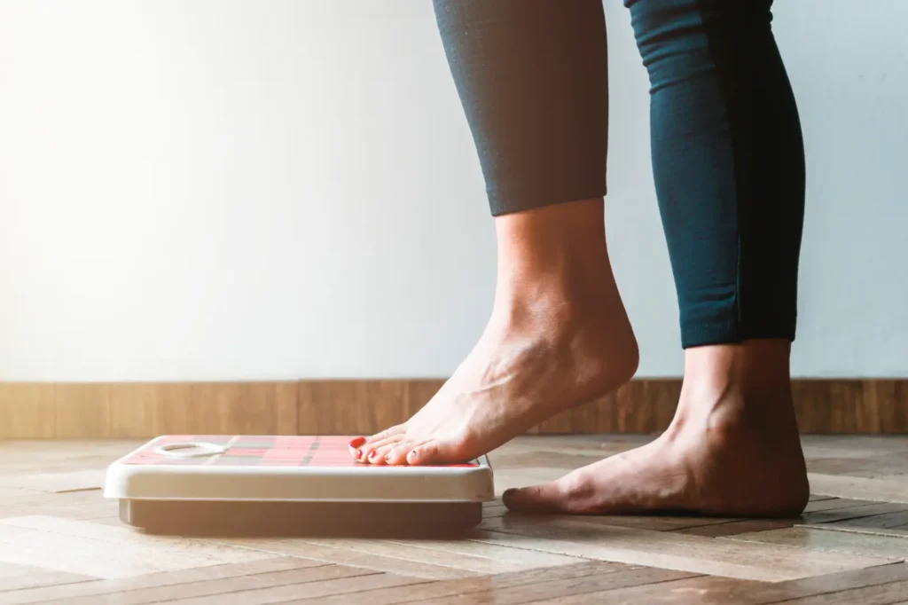 Person stepping onto a bathroom scale with one foot while standing on a wooden floor.